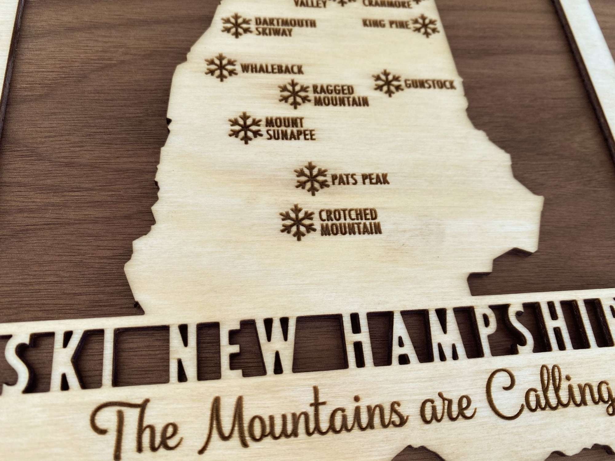 Close-up of Ski New Hampshire Map - Wood Wall Decor - Mountaincut highlighting engraved ski resort names and snowflake icons on light wood.