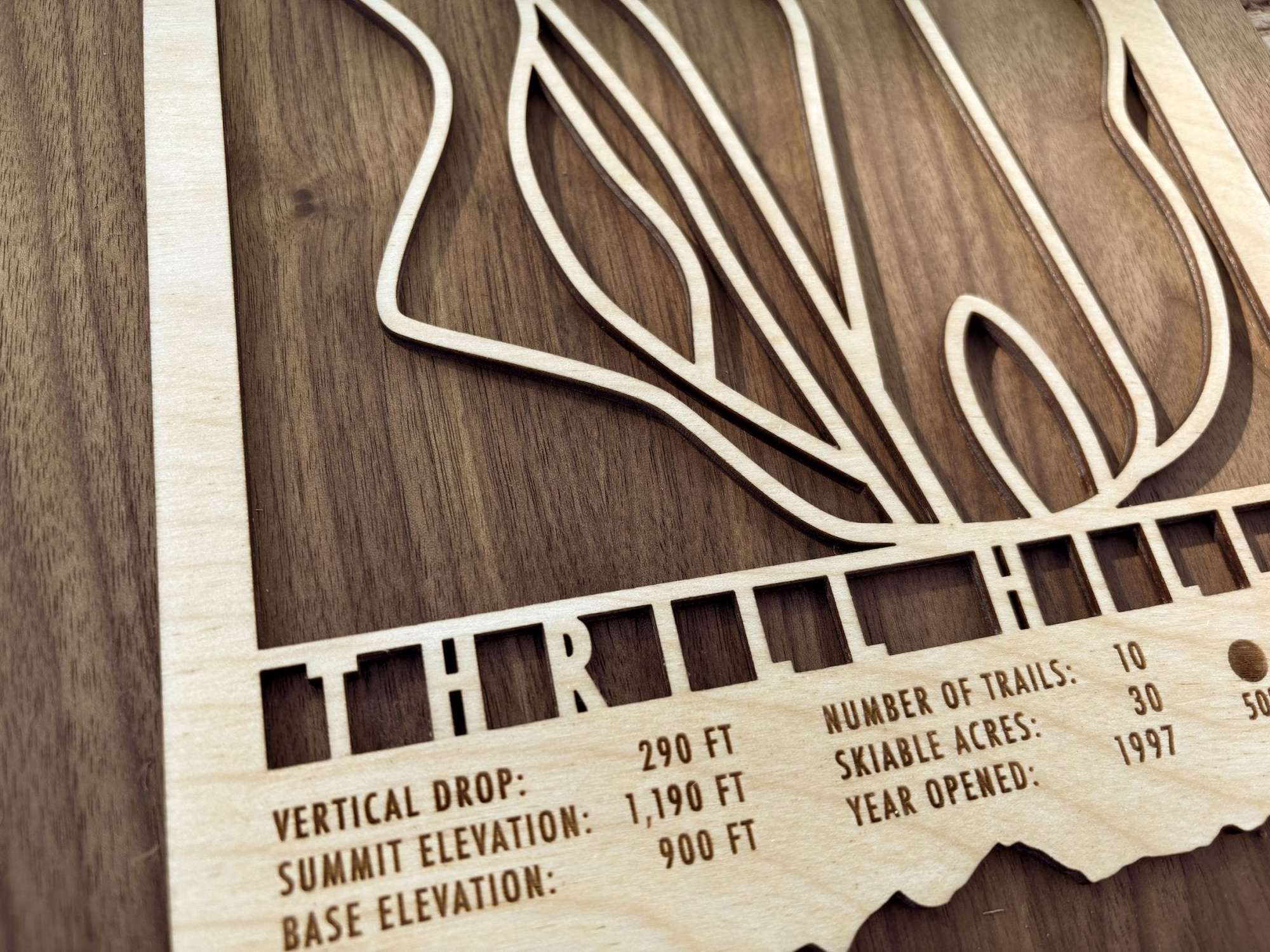 Close-up of Thrill Hills Ski Trail Map showing engraved trail names, vertical drop, and skiable acres on a wood background.