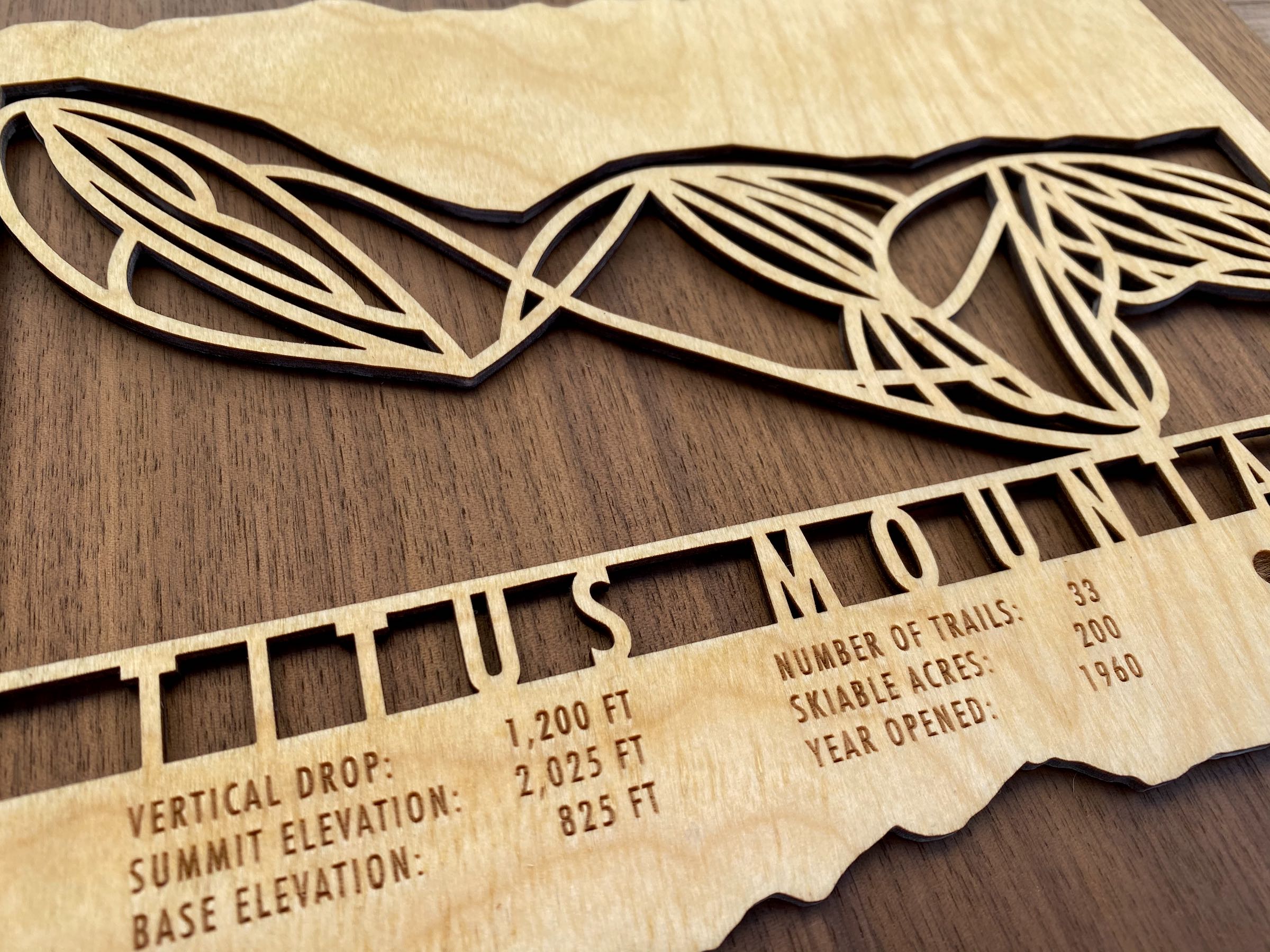 Close-up of Titus Mountain wood ski map showing engraved vertical drop, summit elevation, and number of trails in artistic cutout.